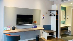 A modern hotel room with a flat-screen TV, a small desk with a chair, a bench, a trash can, and a bathroom with a sink and mirror. - Holiday Inn Express Linyi West by IHG (Linyi)