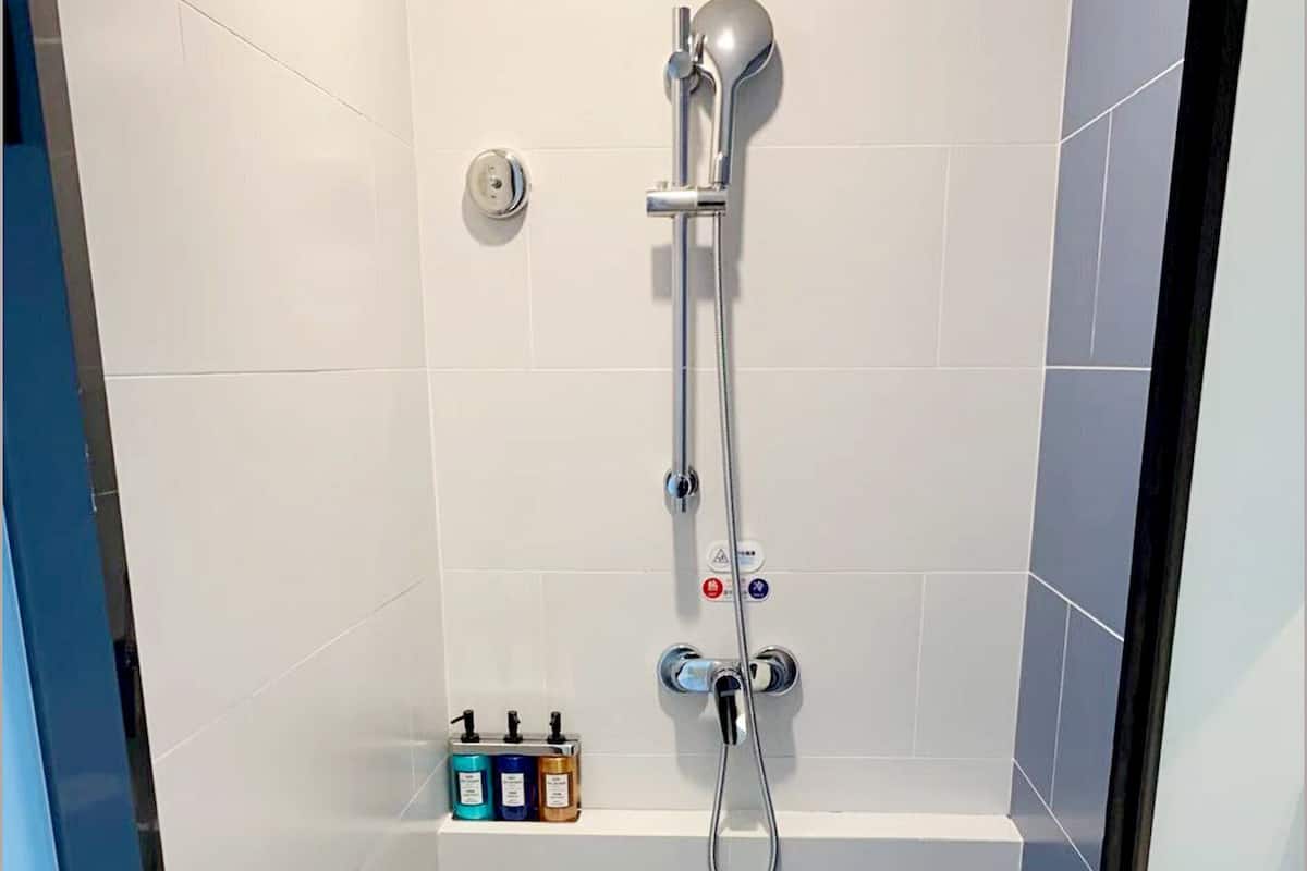 A small, tiled shower area with a handheld showerhead, a control knob, and a shelf with toiletries.