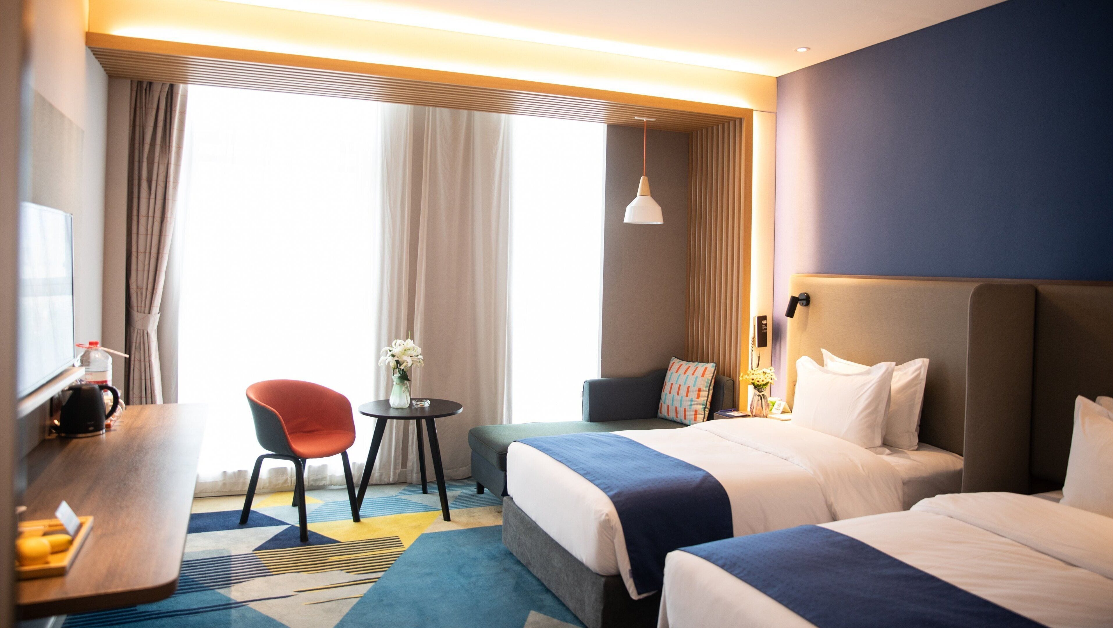 Ein Hotelzimmer mit zwei Betten, einem Schreibtisch, einem Stuhl, einem kleinen Tisch und einem großen Fenster mit Vorhängen.