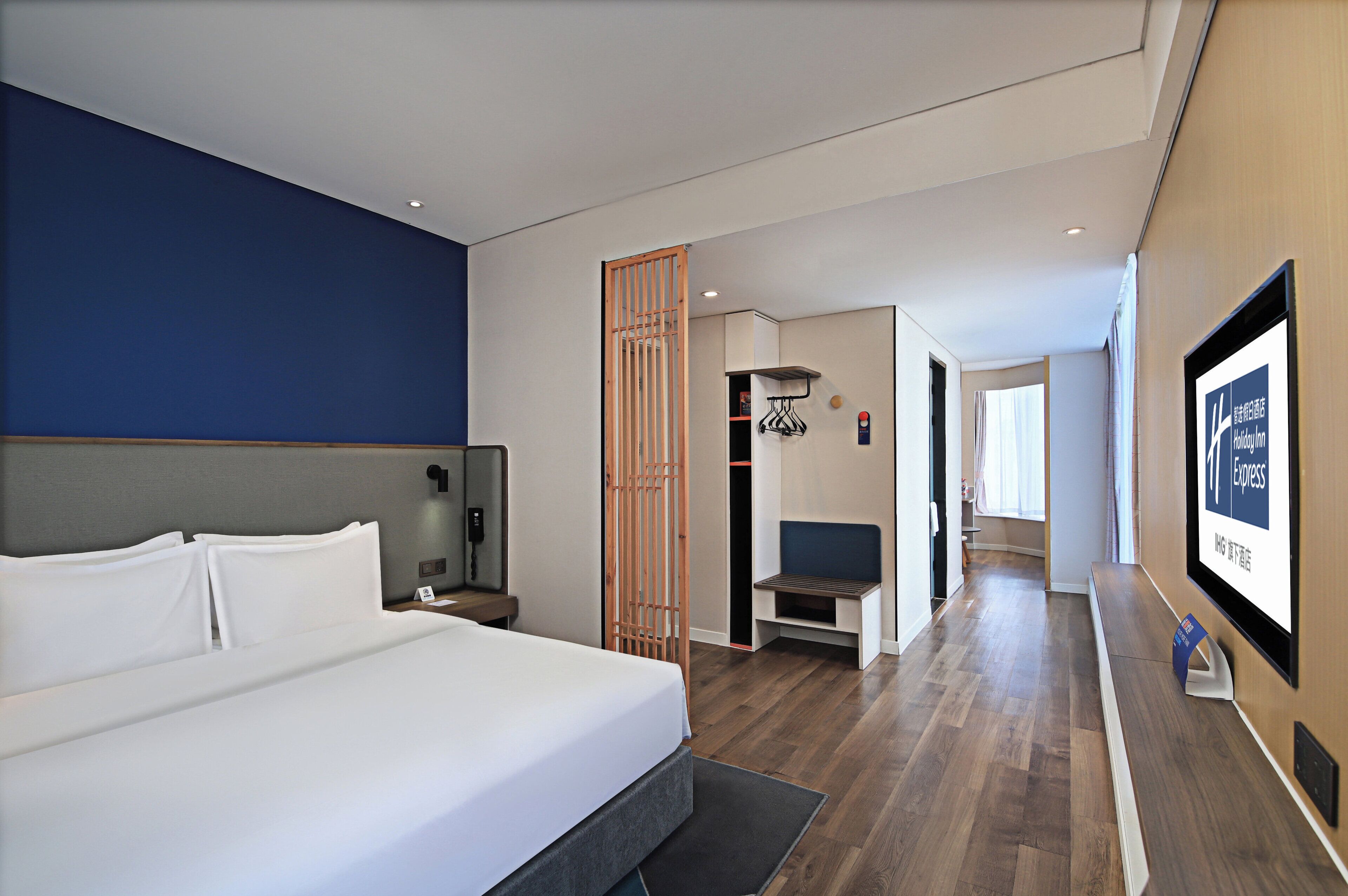 A modern hotel room with a large bed, a desk, a chair, a wooden partition, and a view of the hallway.
