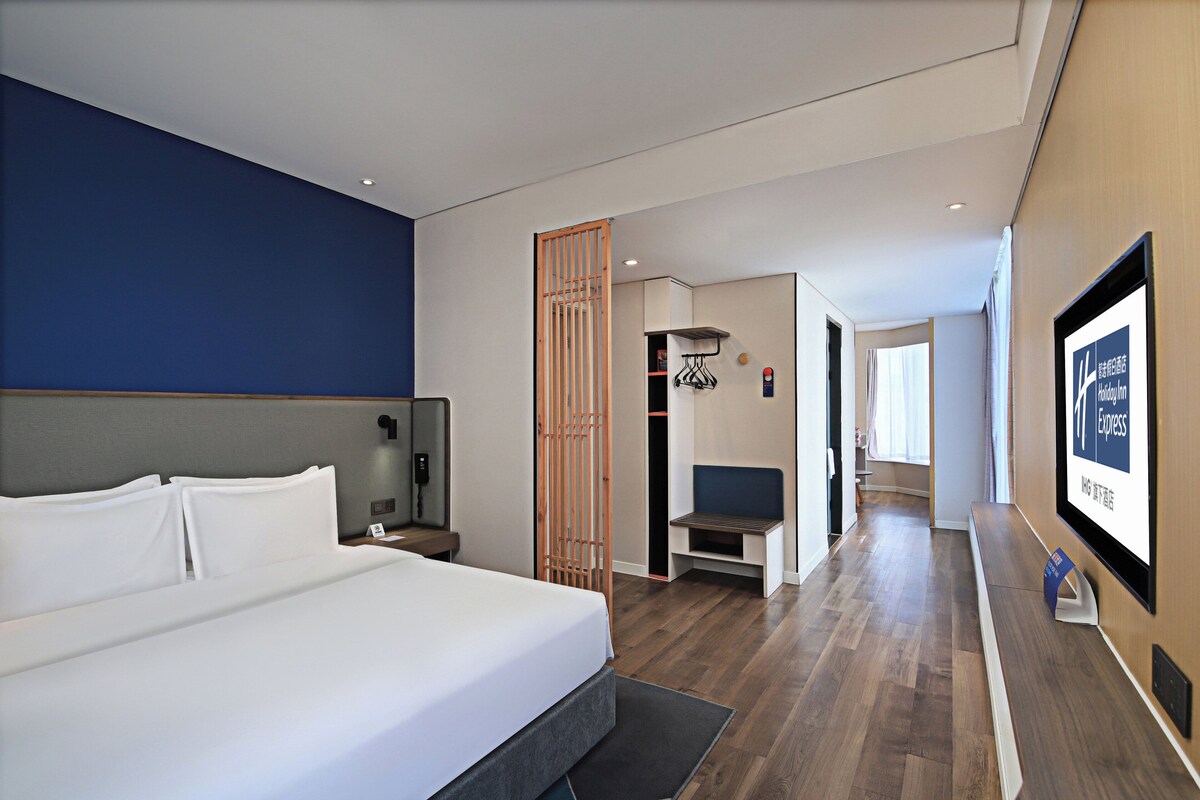A modern hotel room with a large bed, a desk, a chair, a wooden partition, and a view of the hallway.