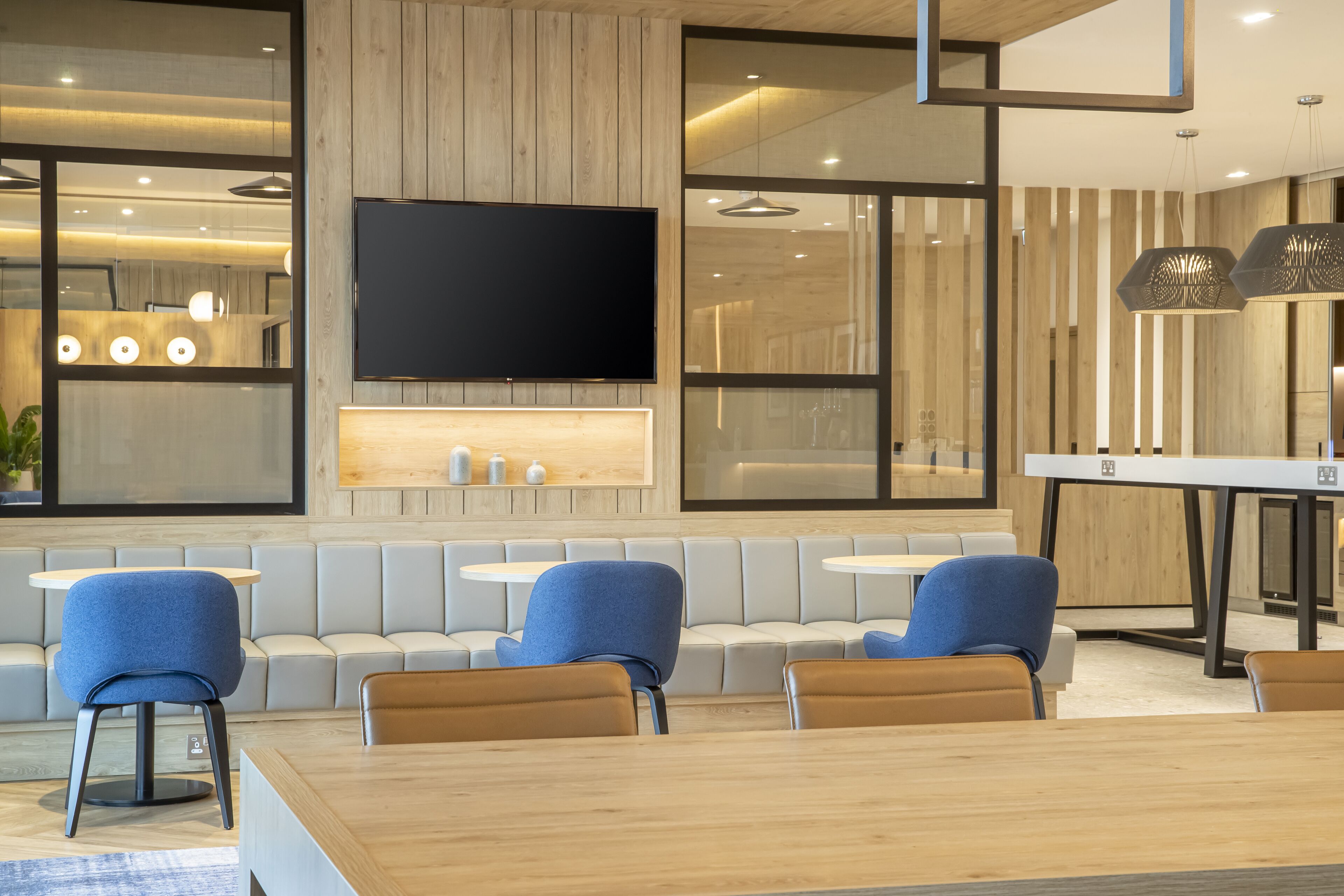 Un interior moderno con un televisor de pantalla plana grande montado en una pared de madera, zonas de estar con sillas azules y marrones, y una mesa de madera.