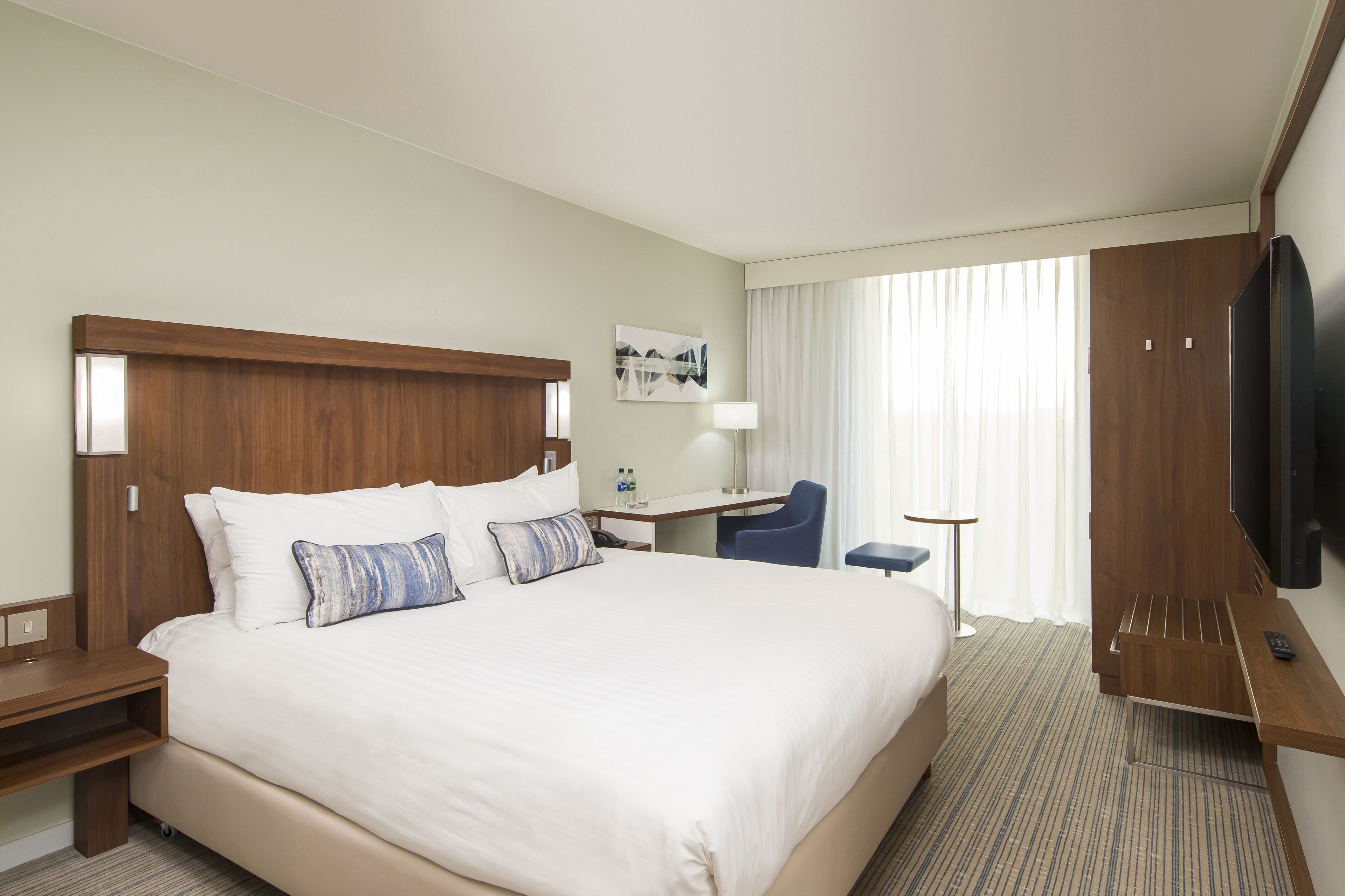 A hotel room with a large bed, a desk, a chair, a TV, and a window with curtains.