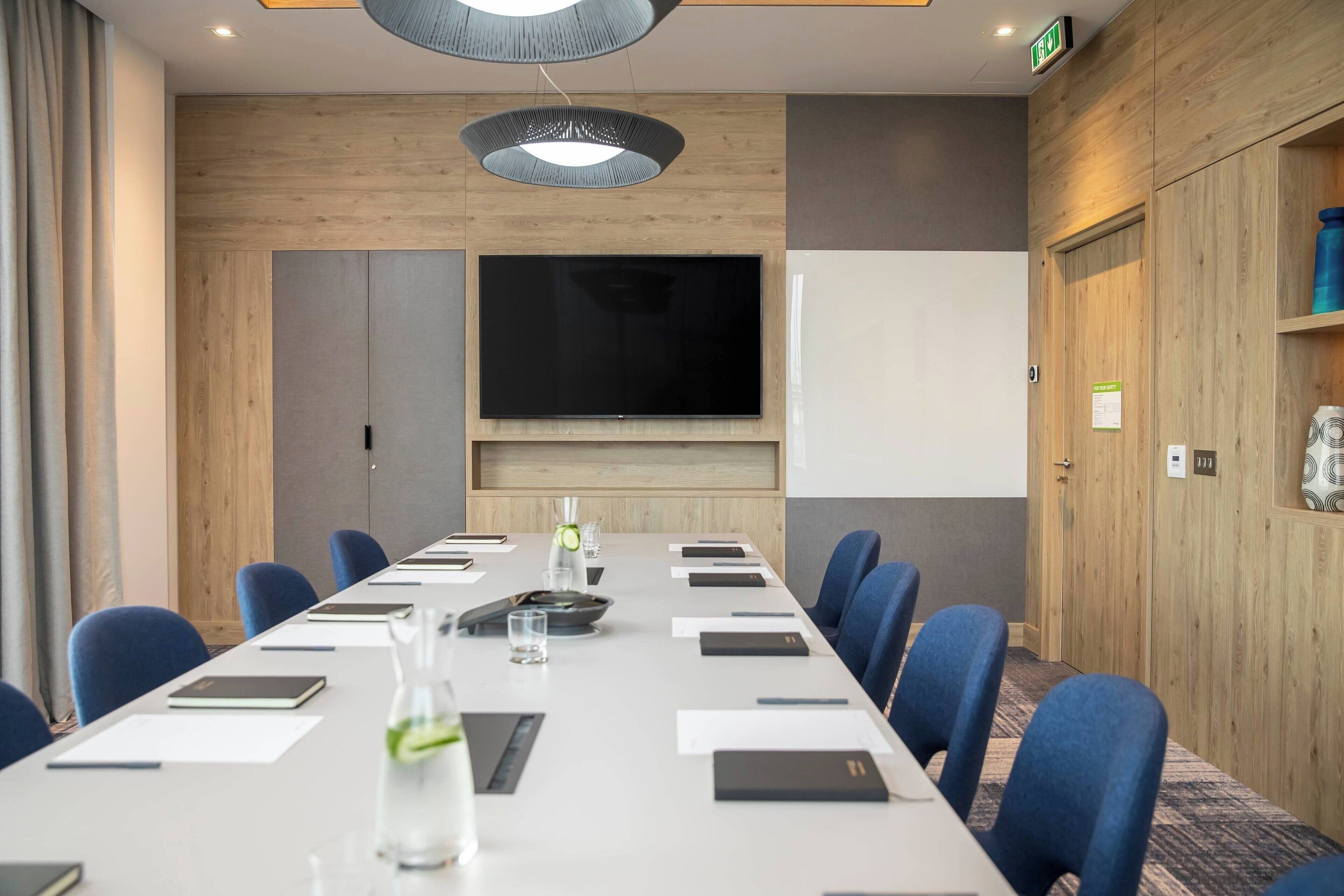 Un salón de conferencias con una larga mesa, sillas azules, un televisor de pantalla plana y un revestimiento de pared de madera.