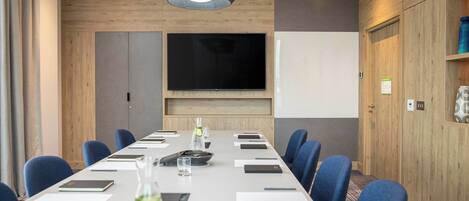 Un salón de conferencias con una larga mesa, sillas azules, un televisor de pantalla plana y un revestimiento de pared de madera.