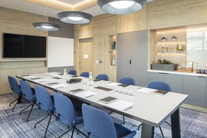 Un salón de conferencias con una larga mesa, sillas azules, una televisión de pantalla plana y lámparas colgantes.