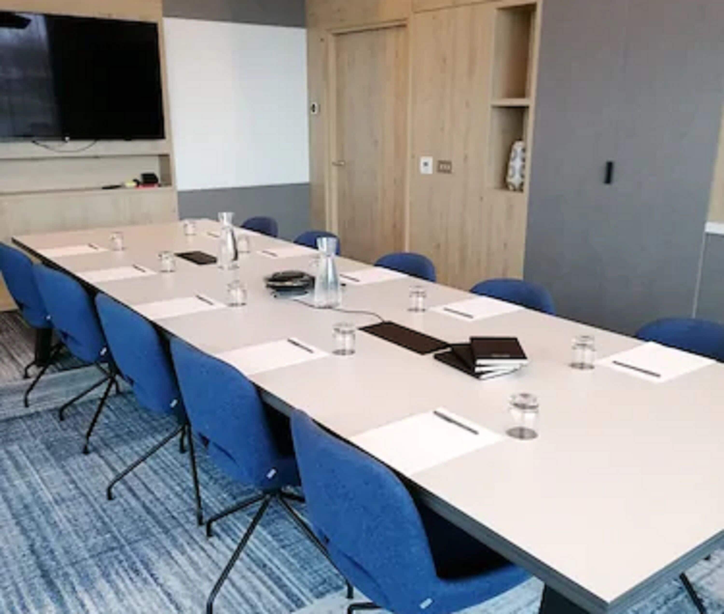 A conference room with a long table, blue chairs, a projector screen, and a wooden cabinet.