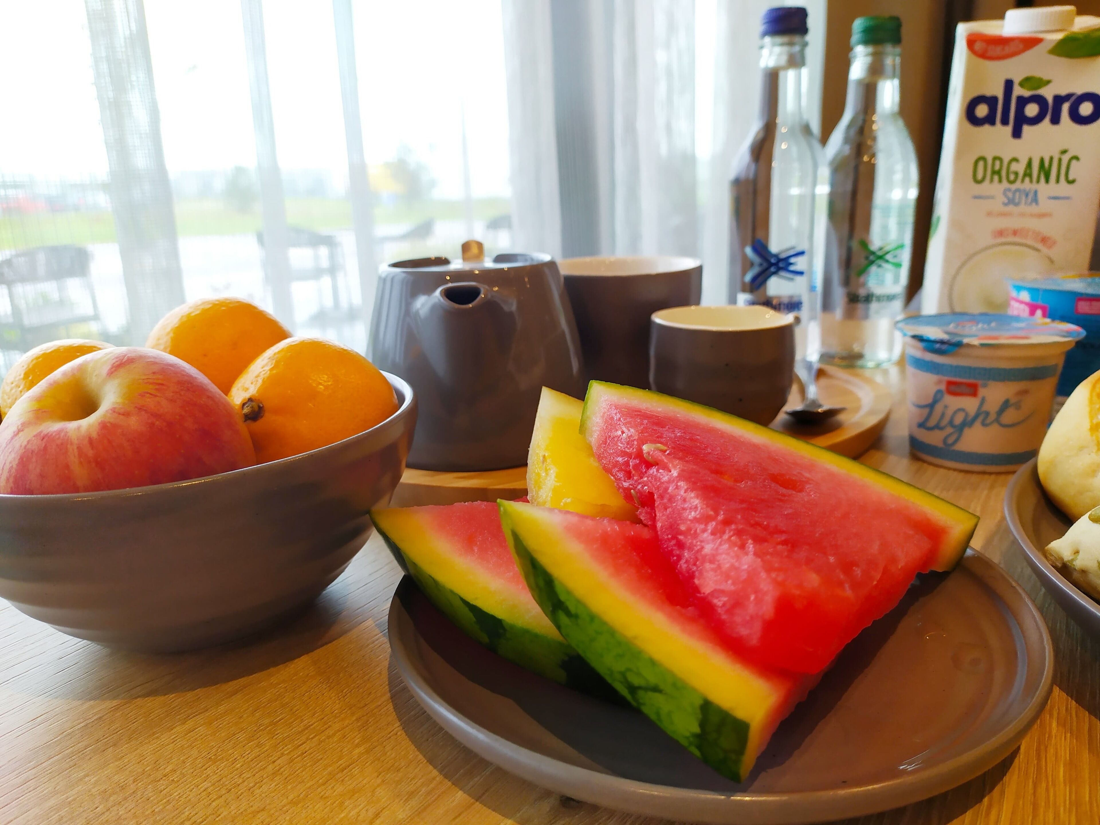 A breakfast table with a bowl of fruit, a teapot, a cup, and a carton of organic soya milk.