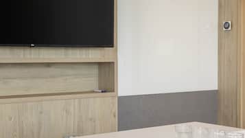 Sala de conferências com uma televisão de tela plana grande presa na parede, uma mesa com caderno, caneta e óculos, e uma cadeira azul.