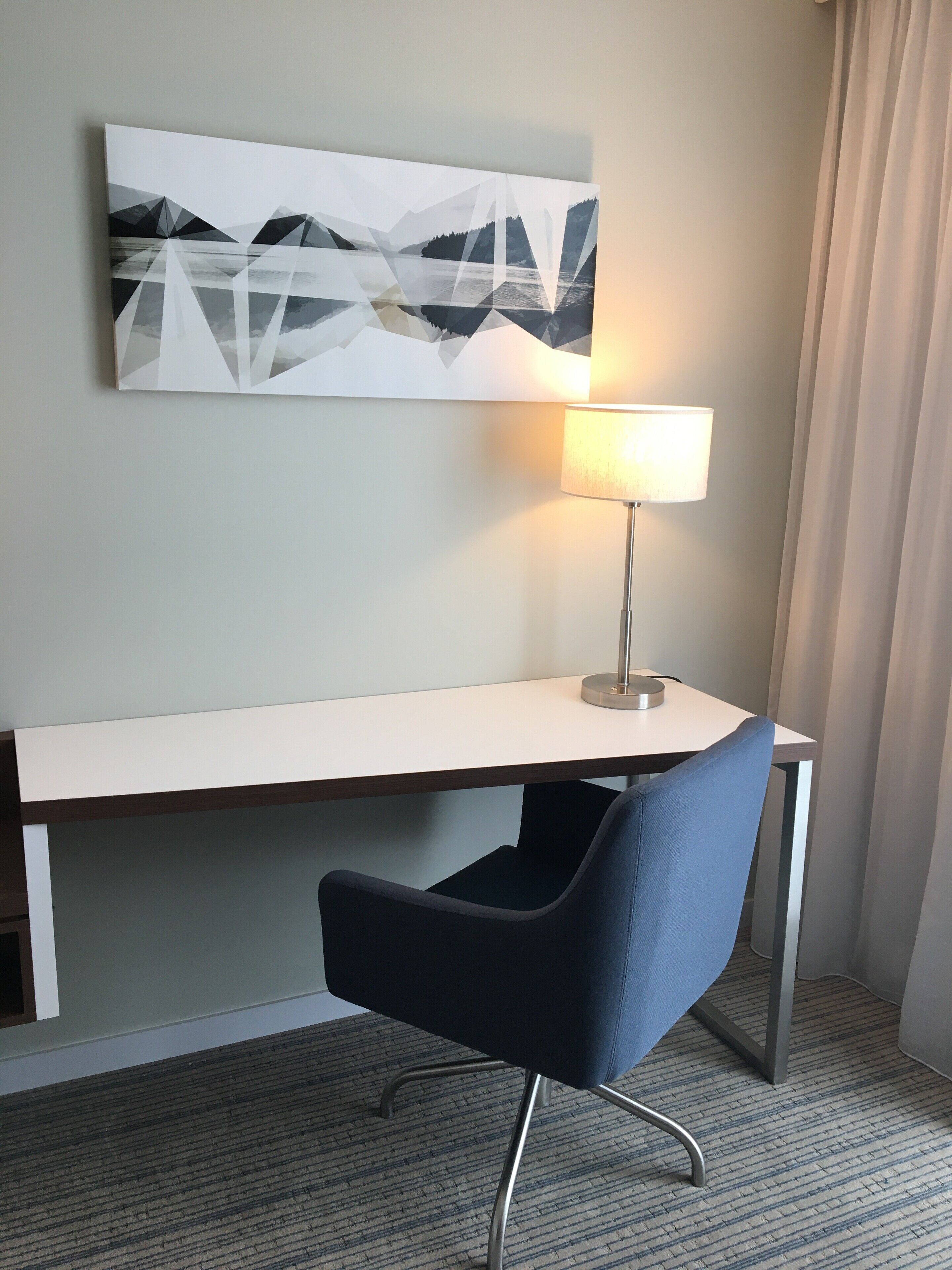 A hotel room with a desk, a chair, a lamp, and a painting on the wall.