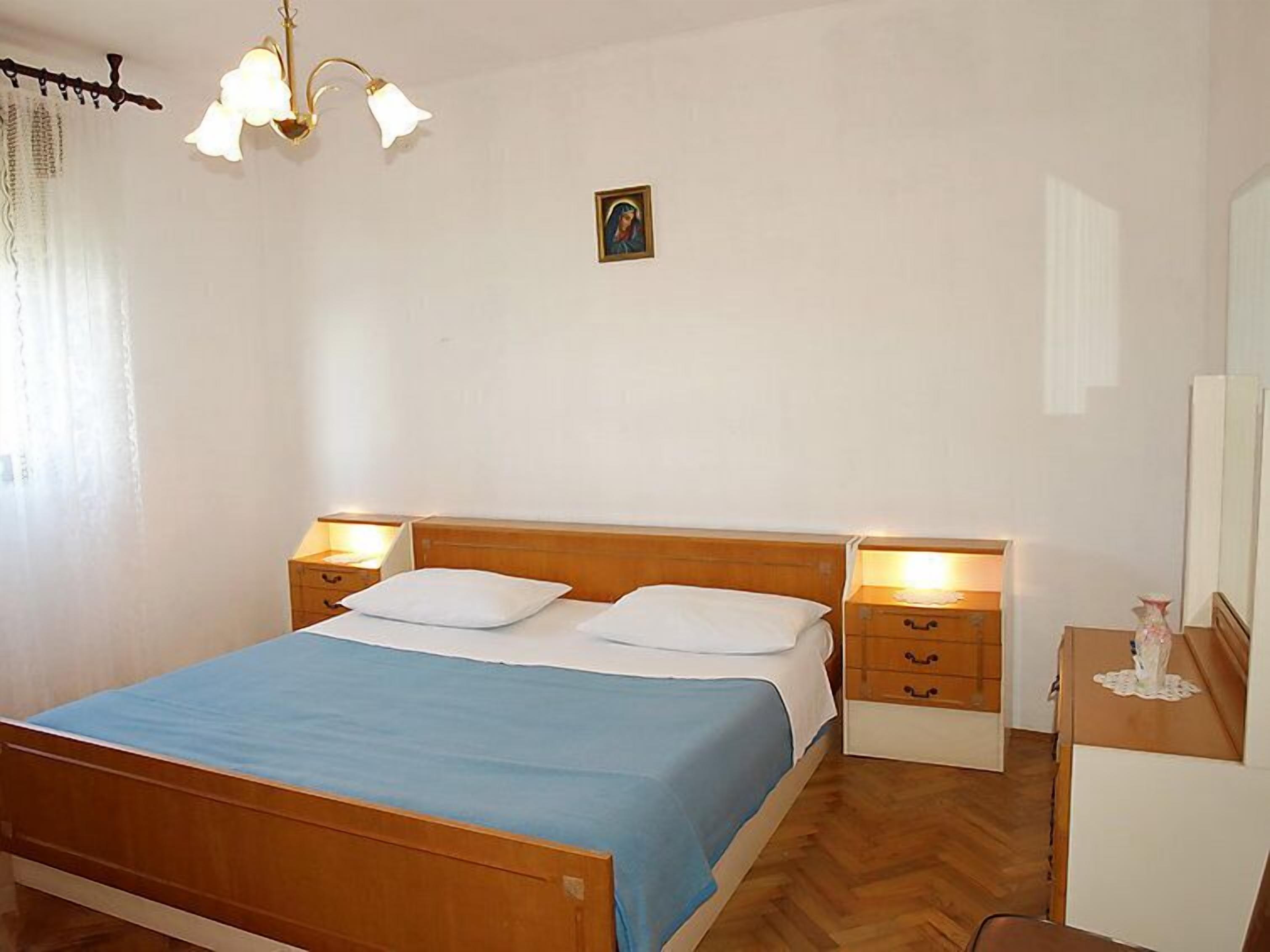 A bedroom with a wooden bed, two bedside tables with lamps, a small bedside table with a vase, and a picture on the wall.