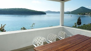 Un balcon avec une table en bois et des chaises en plastique blanches, donnant sur un lac paisible avec des montagnes en arrière-plan.
