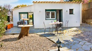 Un petit bâtiment blanc avec un toit en tuiles, une terrasse avec une table et des chaises, et un chemin en pierre.