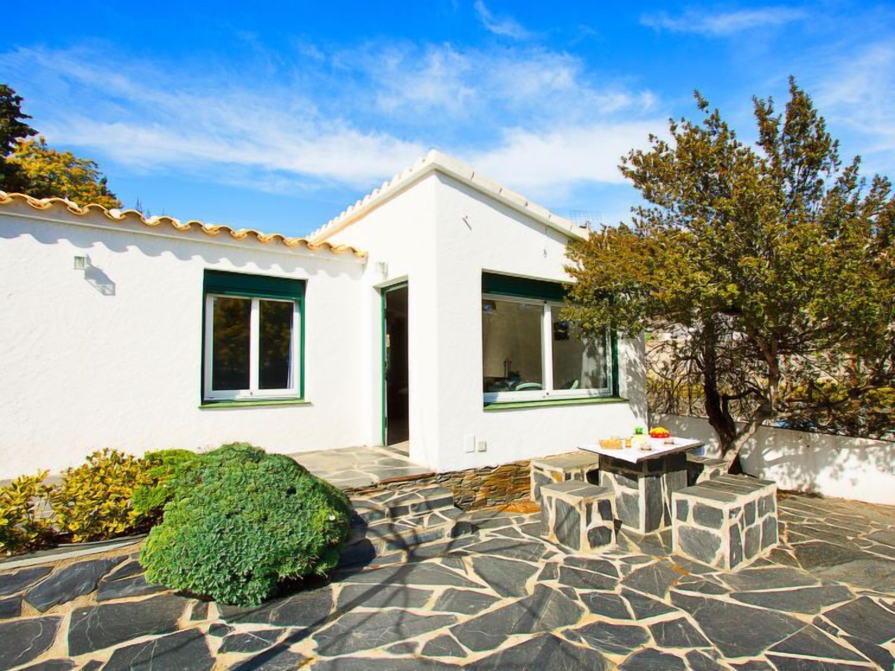 A white building with a tiled patio, a stone table, and a green bush.