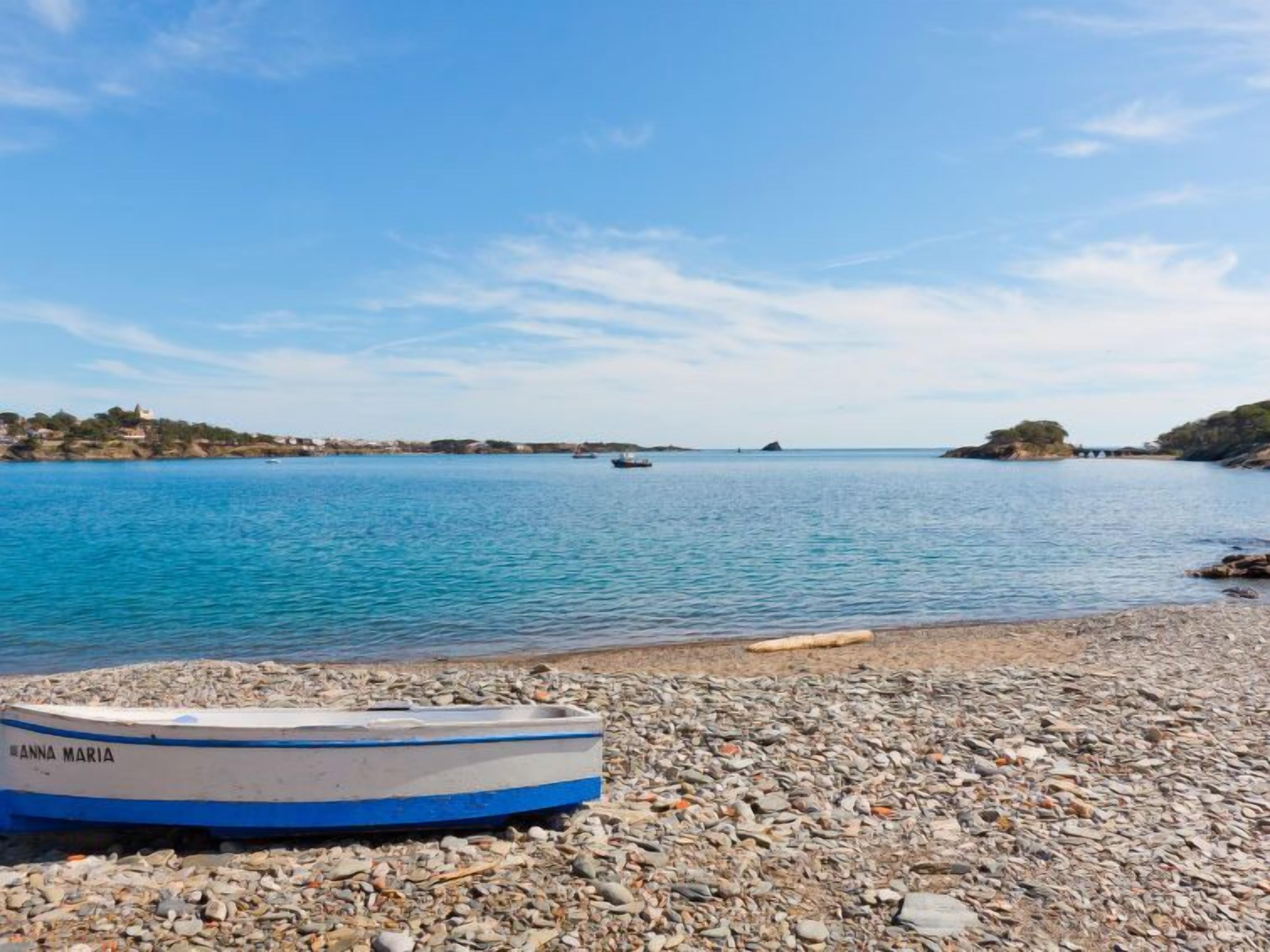 Una playa de guijarros con una lancha llamada "ANNA MARIA" y aguas azules tranquilas.