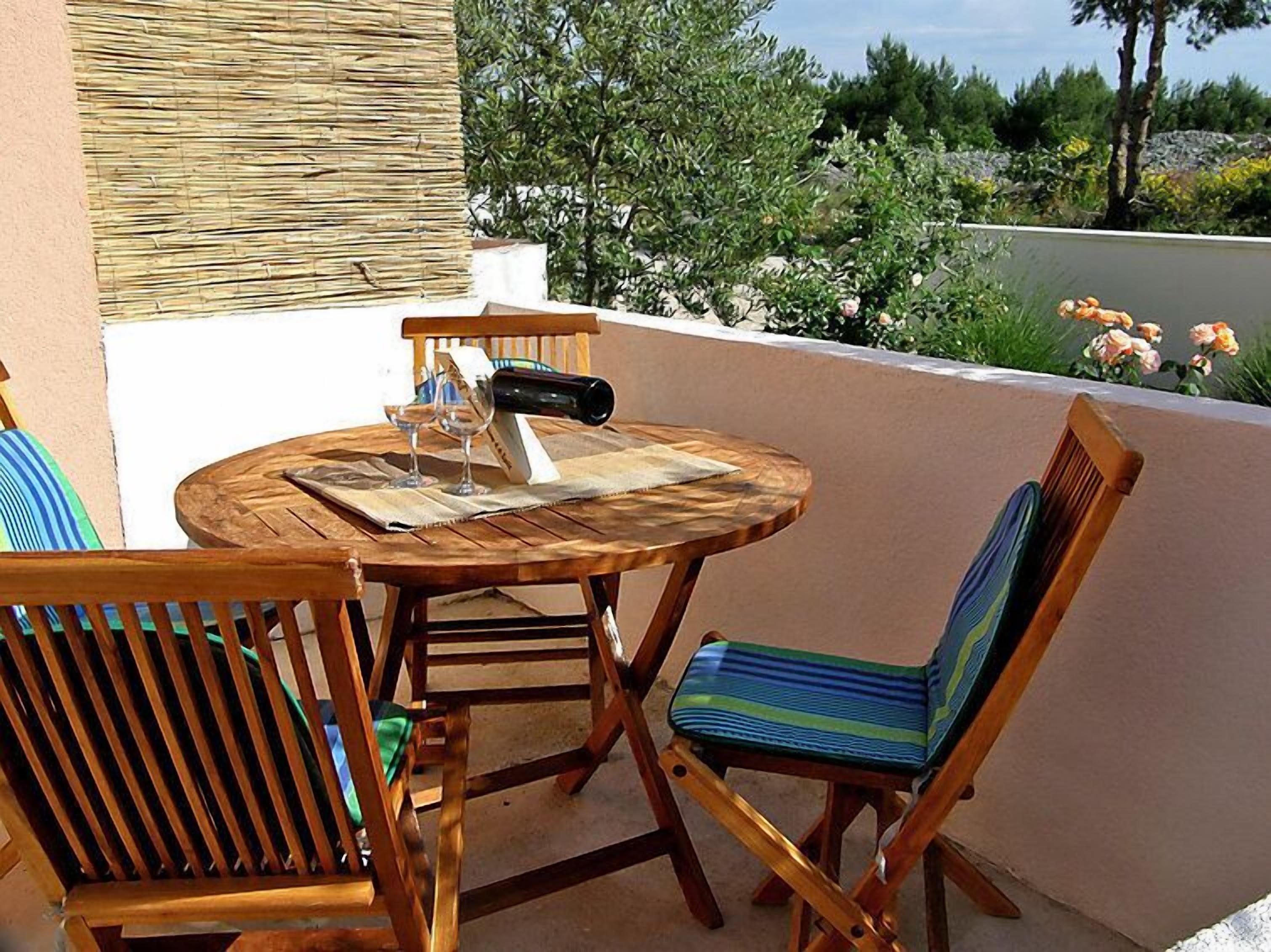A round wooden table with two chairs, a bottle, and glasses on a balcony with a view of greenery.