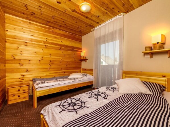 A wooden cabin room with two beds, a nightstand, a window with curtains, and a wall-mounted shelf.