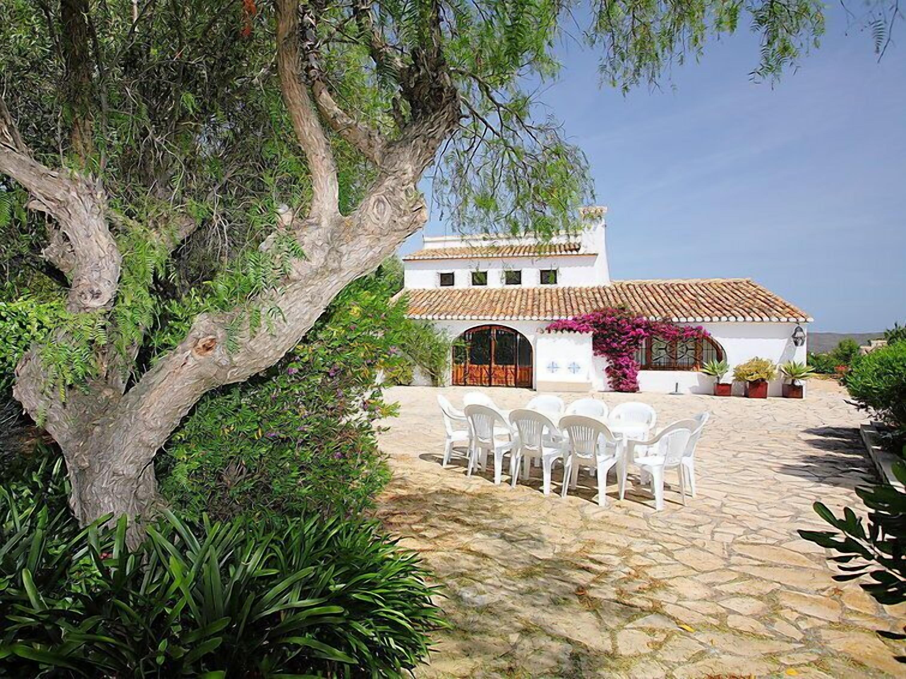 A white building with a tiled roof, surrounded by greenery and a stone patio with white chairs.