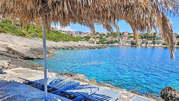 Vista costera con aguas azules cristalinas, un litoral rocoso y una fila de sillas azules para tomar el sol bajo un techo de paja.