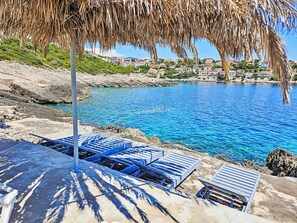 A coastal view with clear blue waters, a rocky shoreline, and a row of blue lounge chairs under a thatched roof. - Snježana by Interhome (Korcula/Zavalatica)