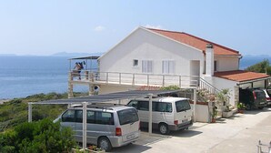 Un edificio de dos pisos con techo de tejas rojas, un balcón y vista al mar. Hay dos furgonetas y un coche estacionados al frente.