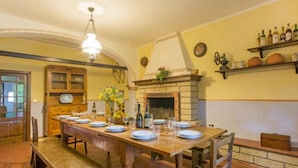 Ein Essbereich mit einem langen Holztisch, gedeckt für sechs Personen, einem Kamin und einem Regal mit Flaschen.