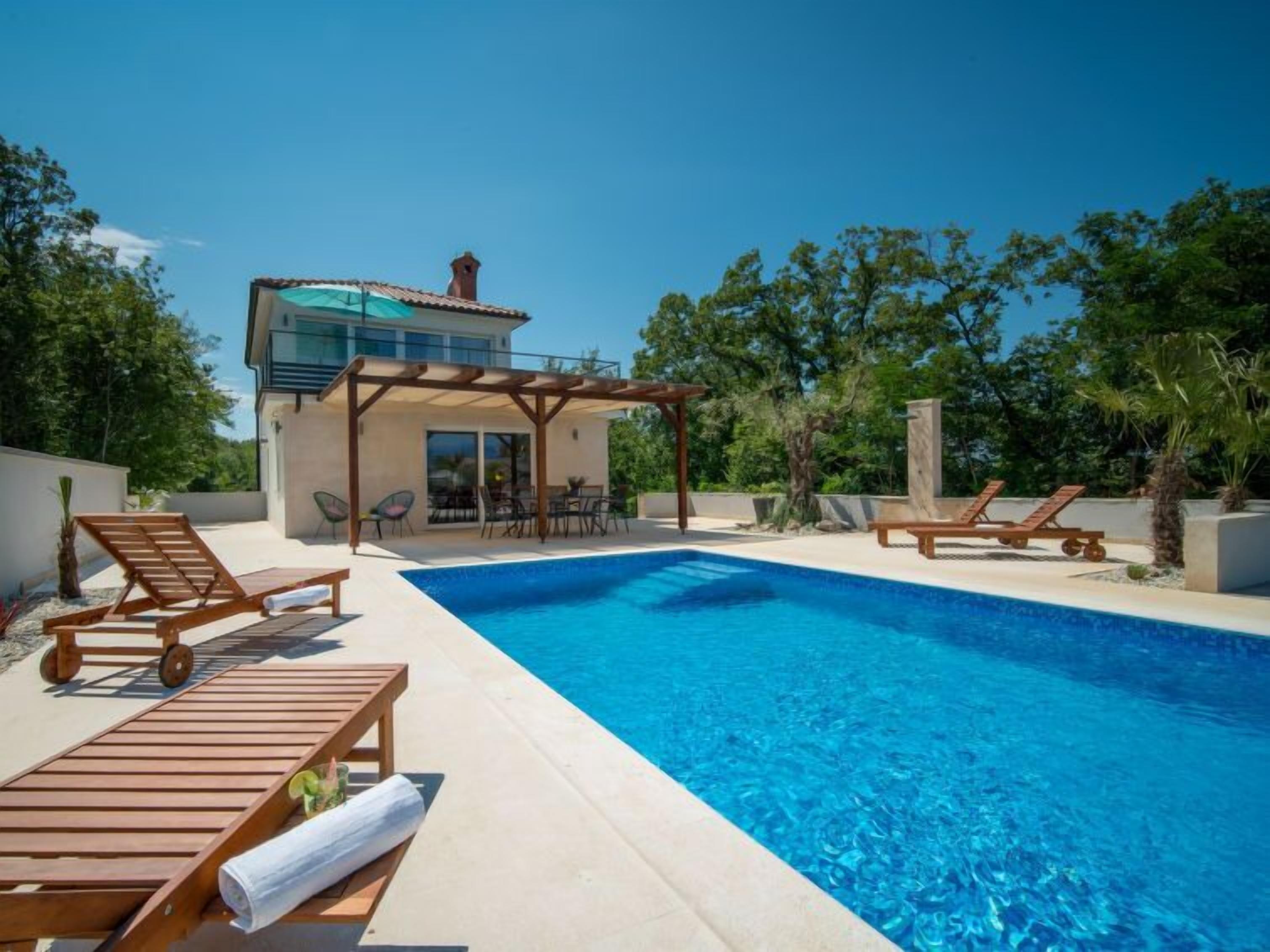 A swimming pool with wooden loungers, a covered patio area, and a modern building with a balcony.