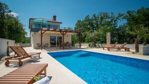 En swimmingpool med træsolstole, en overdækket terrasse og en moderne bygning med altan.