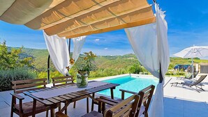 A poolside area with a wooden table and chairs, a white umbrella, and a view of the hills. - Vacation home Ravan by Interhome (Motovun)