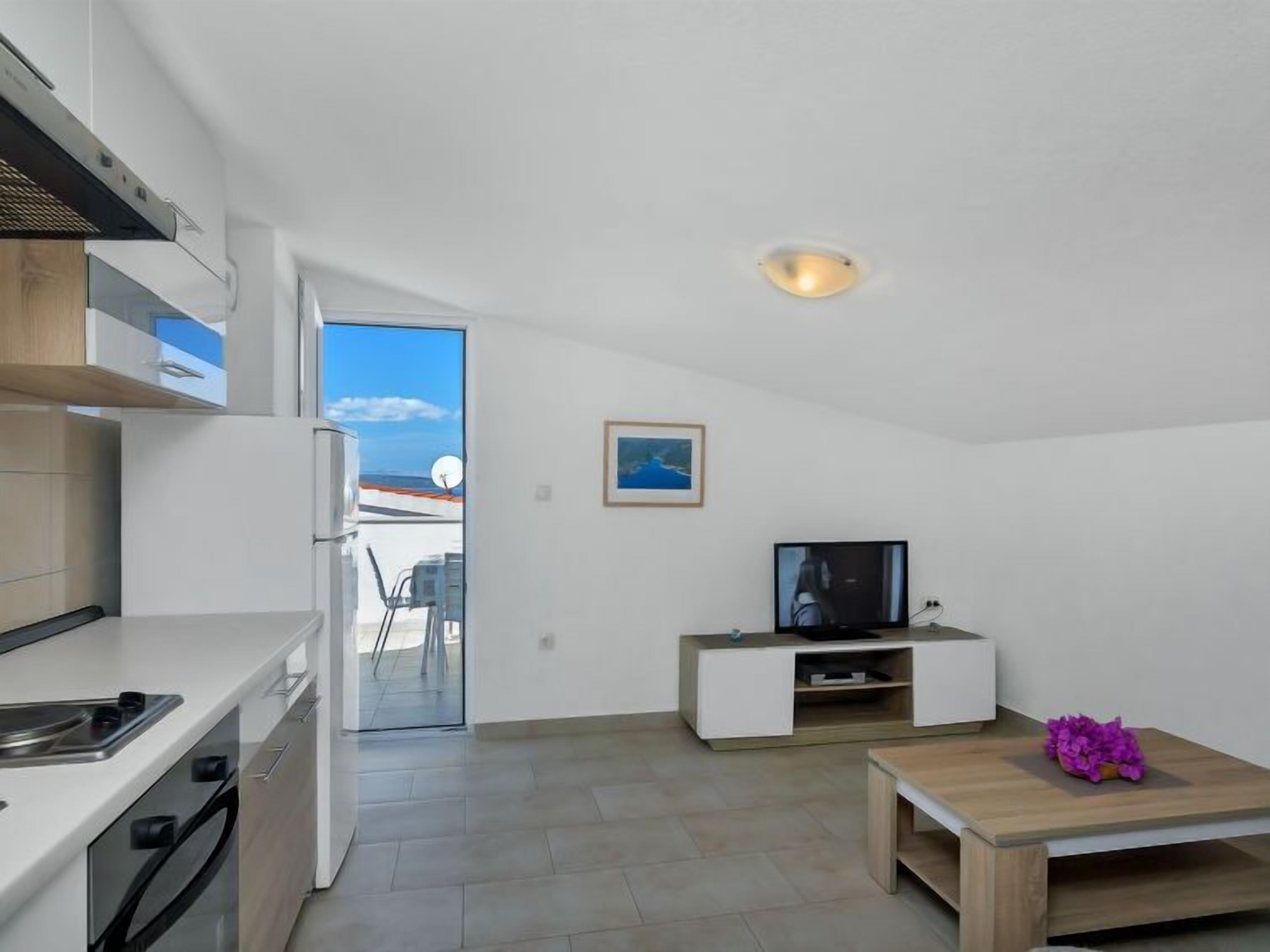 A modern kitchen with a built-in oven and microwave, a dining area with a wooden table and purple flowers, a television on a white stand, and a view of the sea through a glass door.