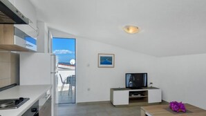 Een moderne keuken met een inbouwoven en magnetron, een eethoek met een houten tafel en paarse bloemen, een televisie op een witte standaard, en uitzicht op zee door een glazen deur.