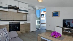 Een moderne keuken met witte kasten, een inbouwoven en een koelkast. Er is een eethoek met een houten tafel en een bank. Een televisie is aan de muur bevestigd, en een raam biedt uitzicht op buiten.