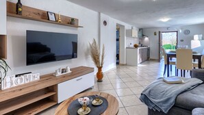 Una sala moderna con televisor de pantalla plana, una mesa de centro y un sofá. Se ve un área de cocina con una mesa de comedor y sillas a través de una puerta abierta.