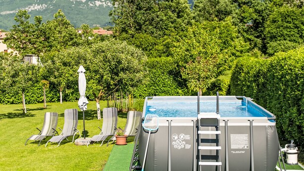 Piscina exterior con escalera, rodeada de sillas de descanso y una planta en maceta, con montañas y árboles al fondo.