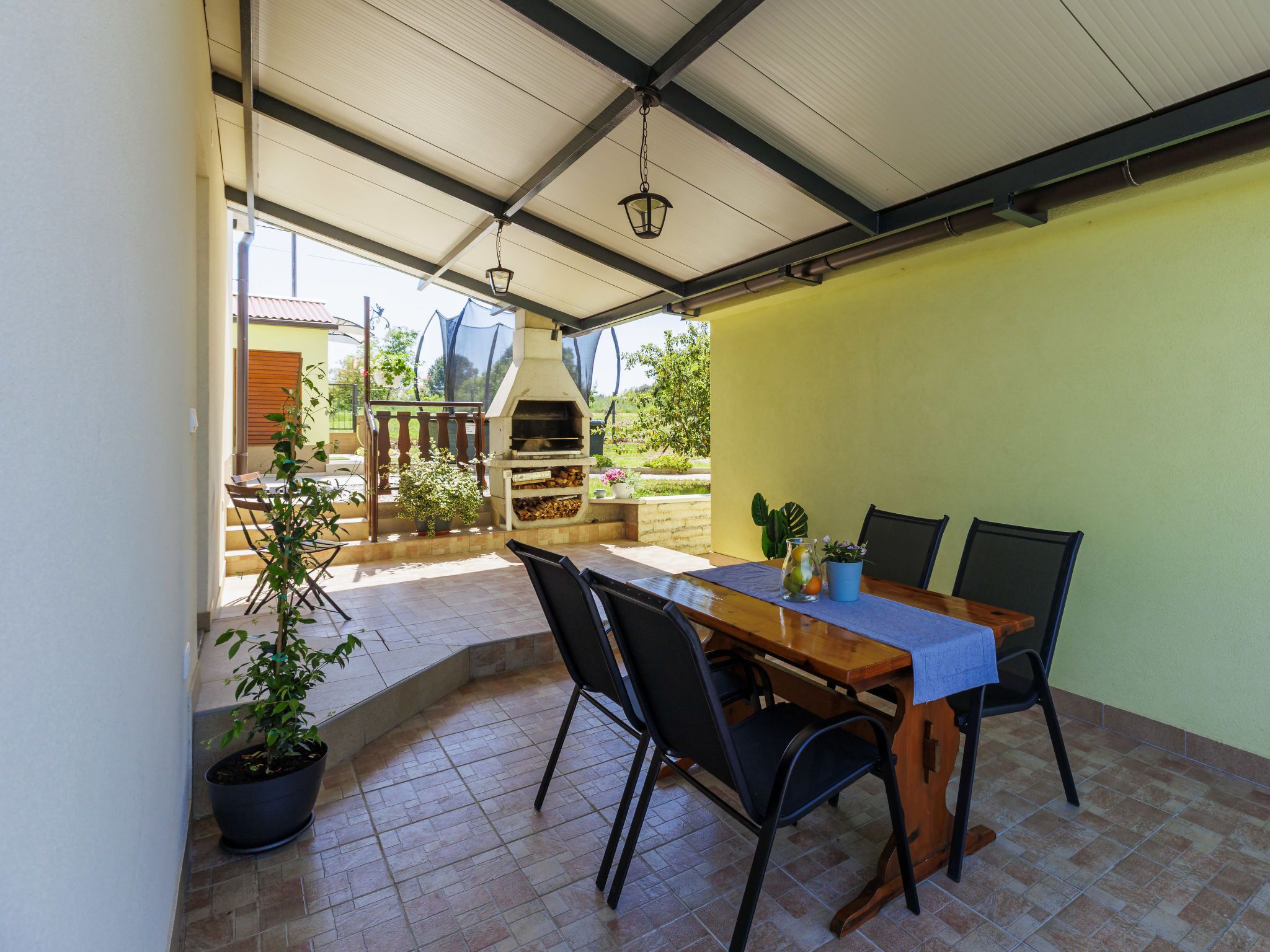 Une terrasse couverte avec une table et des chaises, un pot de fleurs et un barbecue.
