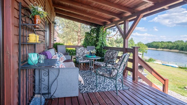 Una terraza cubierta al aire libre con piso de madera, una mesa con sillas y una barandilla con vista a un lago.