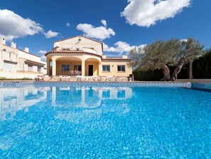 Une piscine avec une eau d’un bleu limpide, un bâtiment doté d’un toit en tuiles et un arbre en arrière-plan.