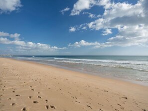 Une vaste plage de sable où l’on peut voir des empreintes de pas menant à l’eau, sous un ciel bleu dégagé parsemé de quelques nuages.