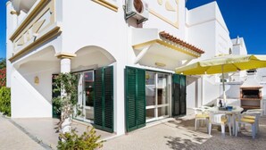 Un bâtiment blanc avec des volets verts, une terrasse dotée de sièges extérieurs et un appareil de climatisation visible.