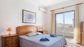 Une chambre à coucher avec un lit en bois, deux tables de chevet avec des lampes, une grande fenêtre avec des rideaux et une œuvre d’art encadrée au mur.