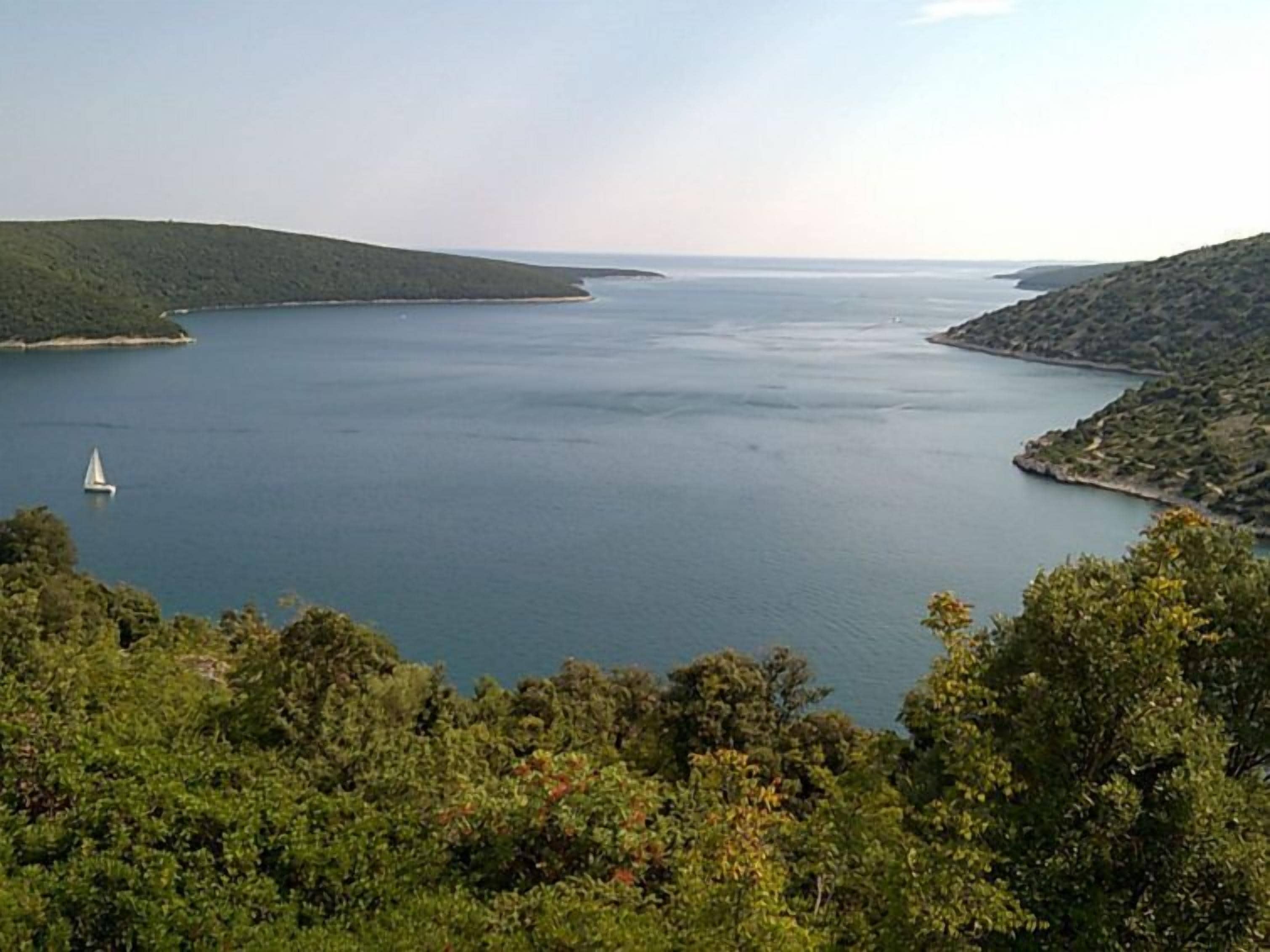 A coastal landscape with a body of water, a sailboat, and surrounding greenery.