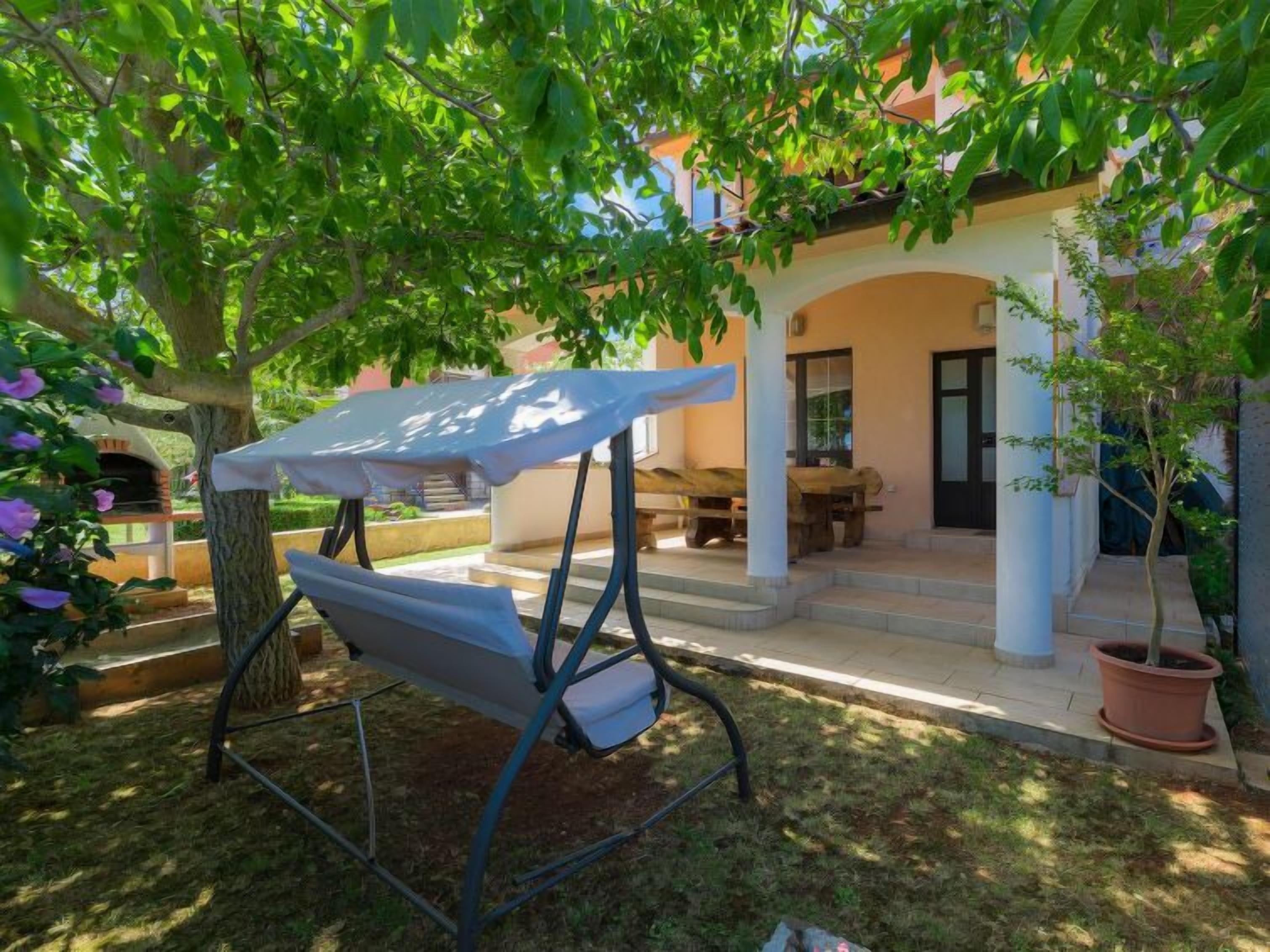 A covered outdoor seating area with a swing chair, a potted plant, and a building with a tiled roof in the background.