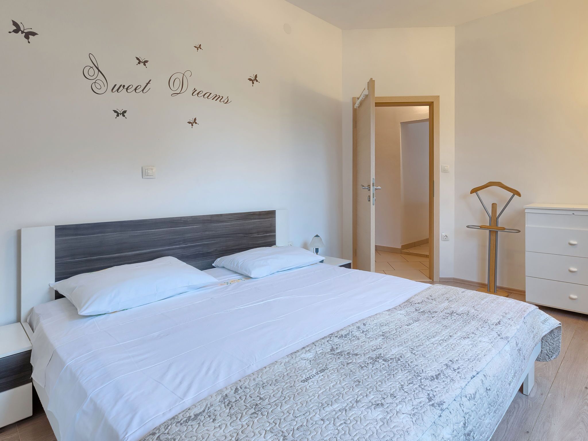 Un lit bien fait, avec du linge de lit blanc, une tête de lit grise et un mur décoré avec les mots « Doux rêves » et des motifs de papillons.