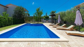 Una piscina rectangular con sillas de descanso y sombrillas, rodeada de un patio empedrado y vegetación.