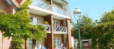 A two-story residential building with a brick facade, a balcony with a glass railing, and a small garden area with a lawn and a swing set.