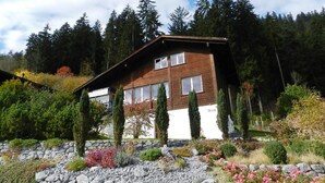 Una casa de dos pisos con un muro de piedra en el frente, rodeada de plantas y árboles.