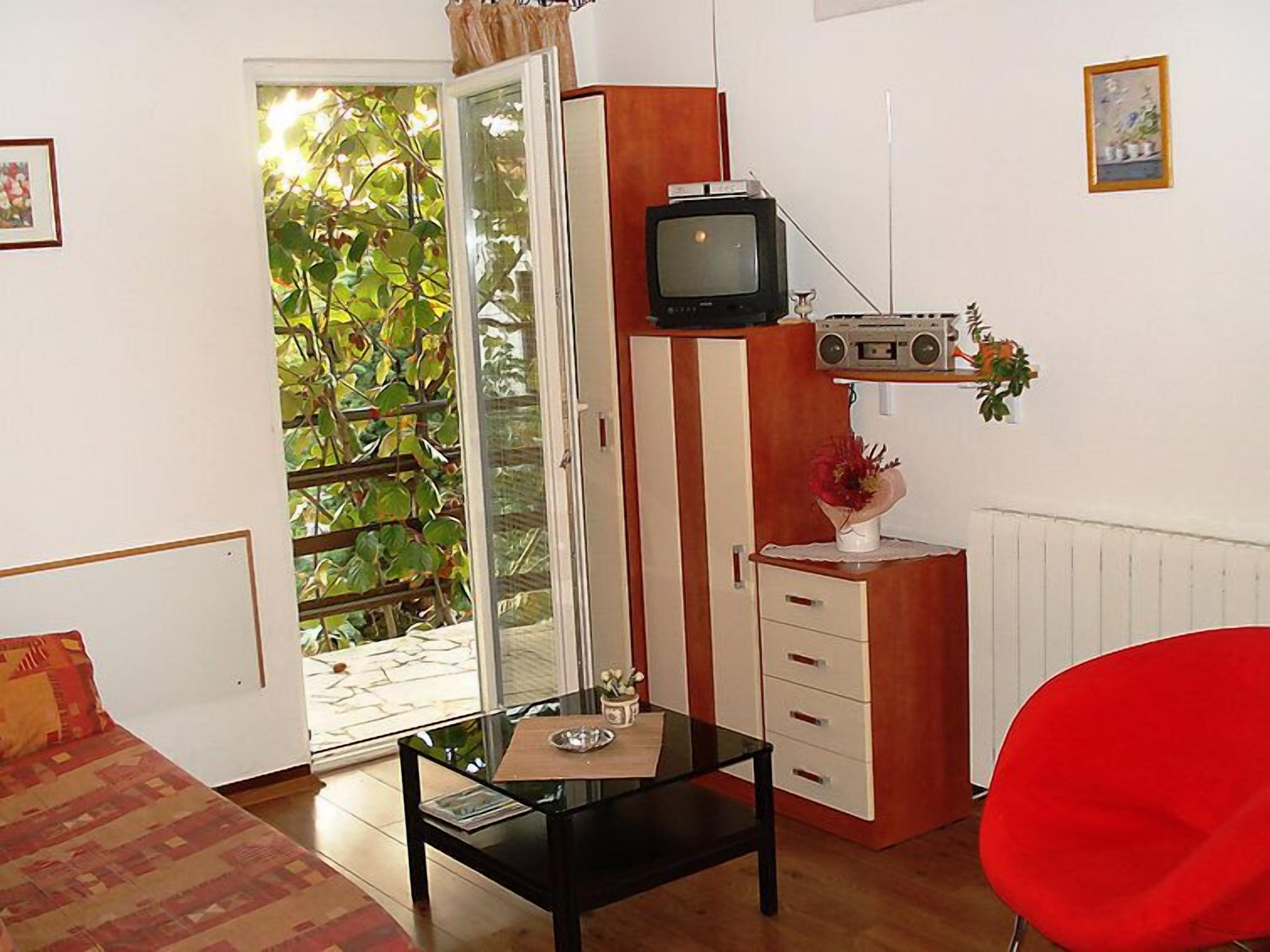 A room with a glass table, a red chair, a wooden cabinet with a radio, and a sliding glass door leading to a balcony.