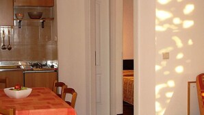 Uma área de refeições com uma mesa posta para dois, uma cozinha ao fundo e uma porta que dá para outro cômodo.