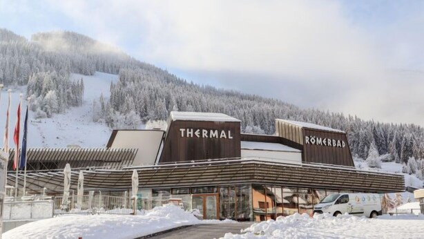 En modern byggnad med skyltar som visar orden ”TERMALBAD” och ”ROMERSKT BAD”, omgiven av snötäckta träd och en bergskedja i bakgrunden.