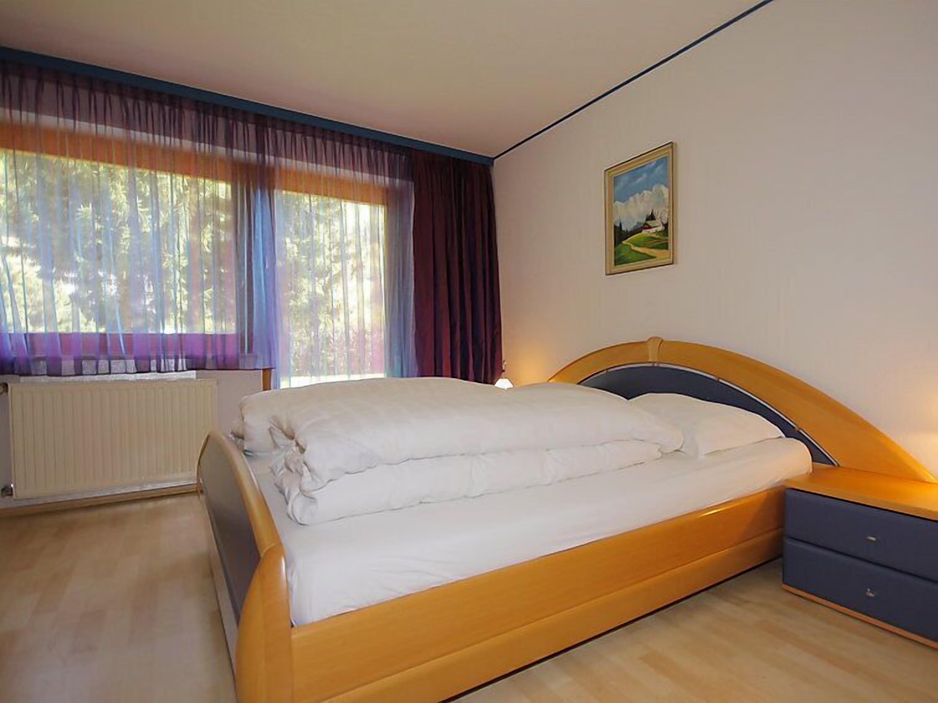 Un dormitorio con cama, mesita de noche, radiador y ventana con cortinas.